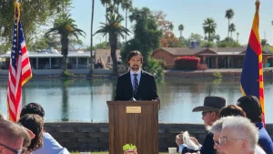 Brad Bettencourt speaking at an event in Arizona in front of a lake and flanked by both an Arizona flag and a US flag